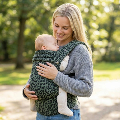 Woman holding a baby in a green leopard print carrier outdoors.
