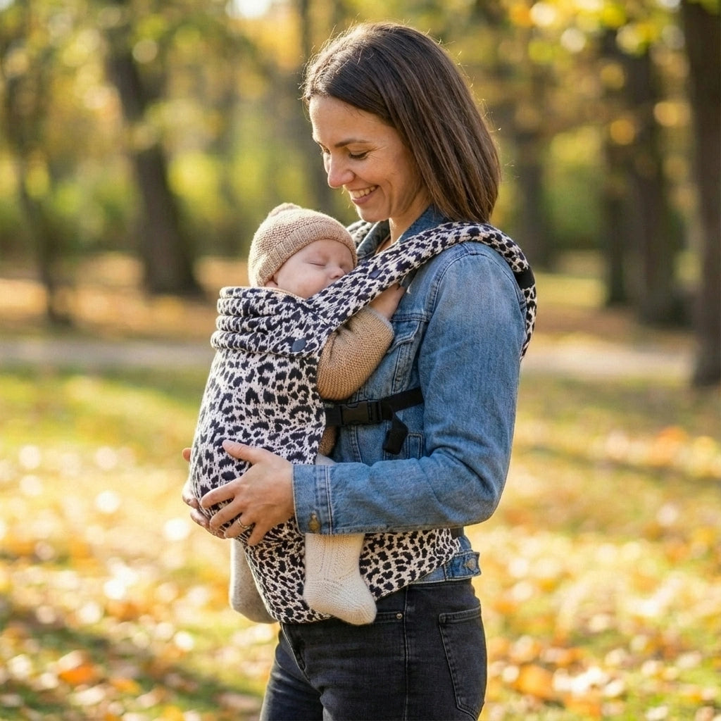 Woman holding a baby in a leopard print wrap outdoors in a park with trees and grass.